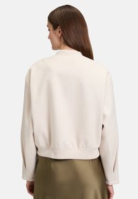 Jasnobeżowa kurtka bomberka z prążkowanym kołnierzem i mankietami, charakteryzująca się miękką fakturą i krótkim krojem. Bez widocznych wzorów ani elementów metalowych.