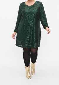 Grön paljettklänning med långa ärmar, rund halsringning och en avslappnad passform, kombinerad med svarta strumpbyxor och gyllene ankeltboots.