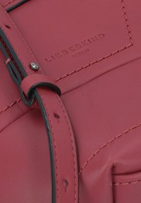 Borsa rossa in pelle con superficie testurizzata, tracolla nera regolabile e logo impresso. I dettagli includono cuciture a contrasto e una chiusura in metallo.