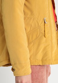 Żółta wodoodporna kurtka z zamkiem z przodu, akcentami z troczkami i kieszeniami po bokach. Gładka tekstura z widocznym wzorem szwów.