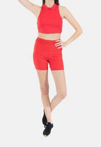 Czerwony crop top i szorty wykonane z elastycznego materiału, z wysokim dekoltem i gładką fakturą. Czarne buty sportowe dopełniają wygląd.
