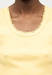 Gros plan d'une personne à la peau foncée portant un débardeur jaune côtelé avec un col rond orné de dentelle et un logo brodé discret.