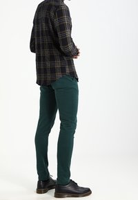 Rutigt skjorta i marinblått, gult och svart, tillsammans med mörkgröna byxor. Svarta skor med texturerad yta och tjocka, bruna sulor.