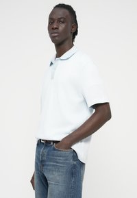 Γαλάζιο πόλο μπλουζάκι με κοντό μανίκι και υφή, με κλασικό γιακά και κουμπιά, συνδυασμένο με μπλε τζιν παντελόνι.