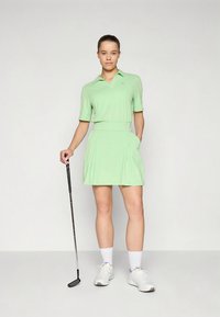 Vaaleanvihreä golfmekko, jossa on kaulus, lyhyet hihat ja laskostettu hame. Kankaan rakenne näyttää sileältä. Yhdistetty golfmailaan ja valkoisiin kenkiin.