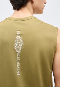 Άνδρας φοράει αμάνικη ελιά πράσινη μπλούζα με λευκή κάθετη επιγραφή "NEVER STOP EXPLORING" και γραφικό σόλας παπουτσιού στην πλάτη.