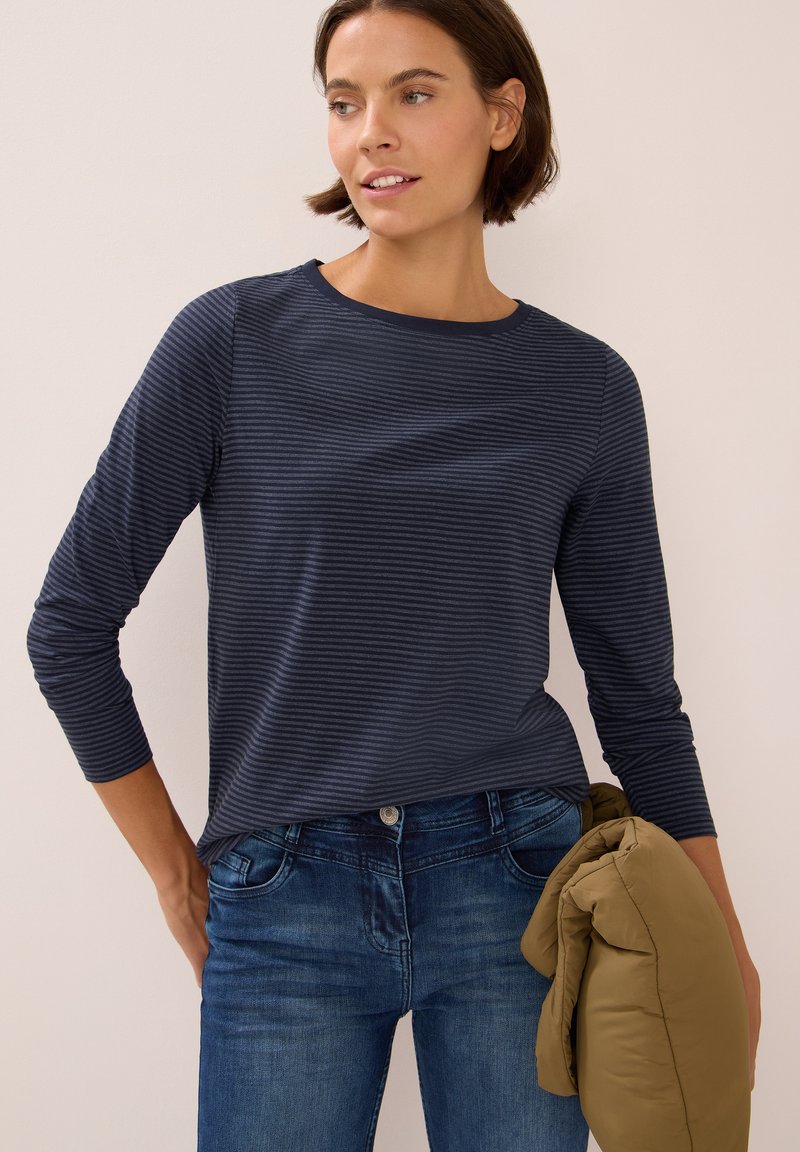 Chemise à rayures marines à manches longues avec un col rond, associée à un jean bleu et un manteau en duvet beige plié tenu dans une main.