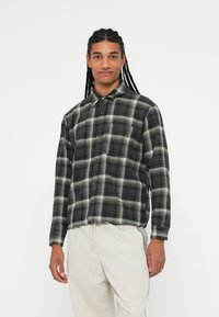 Camicia a maniche lunghe a quadri bianco e nero con colletto button-down e taschino sul petto. Il tessuto appare morbido e strutturato.