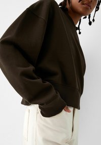 Brun kort sweatshirt med ribbade manschetter, ihop med ljusbeiga byxor. Har en avslappnad passform och rund halsringning.