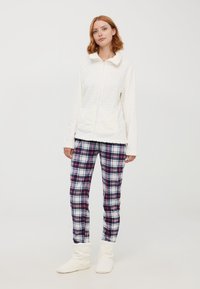 Giacca bianca imbottita con zip e colletto morbido, abbinata a pantaloni a quadri nei toni rosso, blu e bianco, e pantofole bianche pelose.