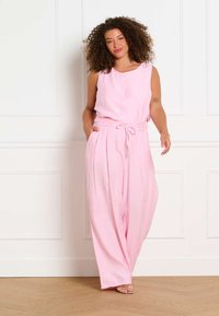 Frau mit lockigem Haar, die einen ärmellosen rosa Jumpsuit mit weitem Bein trägt, steht drinnen vor einer weißen vertäfelten Wand.