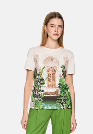 Woman wearing a white t-shirt with a printed design of an arched door, plants, and a leopard lounging by a water pool, paired with green pants.