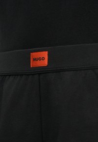 Ceinture en tissu noir avec une petite étiquette rectangulaire rouge affichant le nom de la marque HUGO en lettres noires.