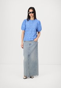 Blouse bleu clair à œillets avec des manches bouffantes, associée à une longue jupe en denim. Des lunettes de soleil noires et des ballerines à bout pointu complètent la tenue.