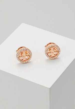 Boucles d'oreilles en or rose avec un design rond, dotées d'un centre en logo sculpté entouré de strass transparents, surface métallique brillante et texture lisse.