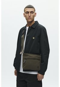 Oliiviroheline kullerikott luku tasku, reguleeritava rihma ja väikese logodetailiga. Kangas tundub vastupidav ja sileda tekstuuriga.