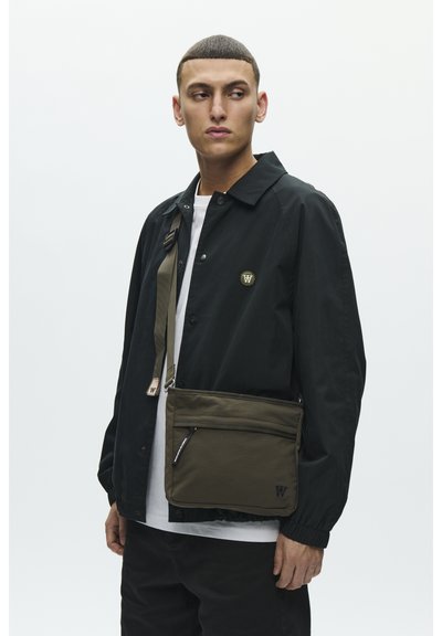 Sac messager vert olive avec une poche zippée, une bandoulière réglable et un petit détail de logo. Le tissu semble durable avec une texture lisse.