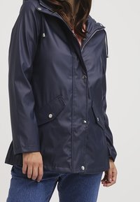 Kvinna klädd i en marinblå vattentålig jacka med tryckknappar och fickor med klaff, ihopkopplad med blå jeans.