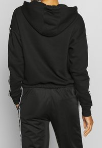 Sudadera negra con una gran capucha, fabricada en tela suave; cuenta con rayas laterales blancas y pantalones de chándal negros de corte relajado con cintura elástica.
