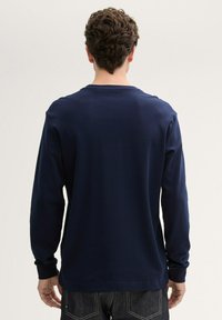 Dunkelblaues Langarmshirt aus einem glatten, weichen Stoff mit Rundhalsausschnitt und gerippten Bündchen, kombiniert mit dunklen Jeans. Keine sichtbaren Muster oder Logos.