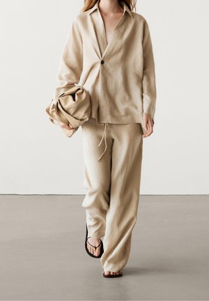 Woman walking in beige loose shirt and pants, holding a matching beige bag, wearing black flip-flops, against a plain light background.