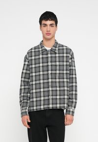 Nuori mies yllään musta-harmaa-valkoinen ruudullinen nappipaita ja mustat housut, seisoo yksinkertaista valkoista taustaa vasten.