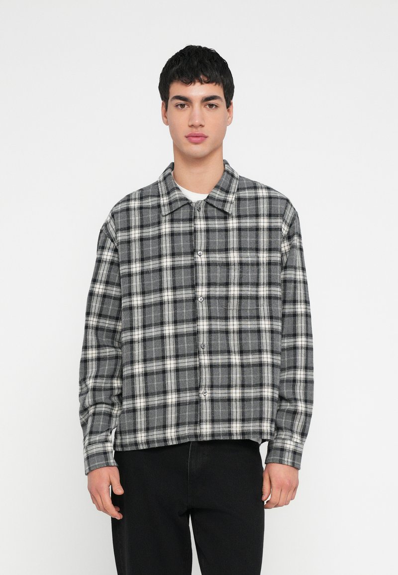 Nuori mies yllään musta-harmaa-valkoinen ruudullinen nappipaita ja mustat housut, seisoo yksinkertaista valkoista taustaa vasten.