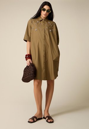 Frau trägt olivfarbenes knopfgeschlossenes Kleid mit Blumenstickerei, grüne Sonnenbrille, rote Armreifen, hält eine braune geflochtene Handtasche und schwarze Slide-Sandalen.