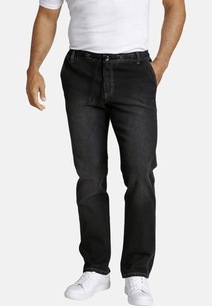 Homme portant un jean noir droit avec un cordon de serrage et des baskets blanches, debout avec une main dans la poche sur un fond uni.