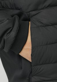 Chaqueta acolchada negra con un tejido texturizado que presenta un patrón de cuadrícula y un puño elástico en la manga. Mano metida en un bolsillo lateral.