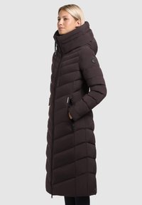 Vrouw in een lange zwarte gewatteerde jas met een hoge kraag, capuchon en ritszakken aan de zijkanten, staand tegen een effen achtergrond.