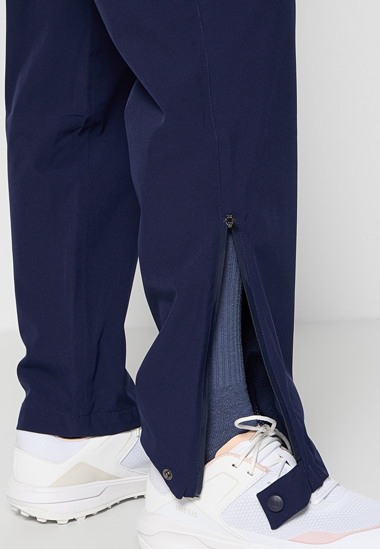 Marineblaue Sporthosen mit seitlichem Reißverschluss, aus einem glatten synthetischen Material. Getragen mit weißen Turnschuhen und einer sichtbaren blauen Socke.