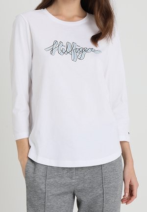Weißes Langarm-T-Shirt mit blauem gesticktem "Hilfiger"-Logo, aus weicher Baumwolle mit rundem Halsausschnitt und lockerer Passform. Graue Hosen sichtbar.