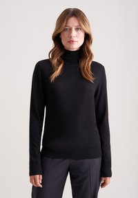 Czarny sweter z wysokim kołnierzem, wykonany z gładkiego materiału, z długimi rękawami i dopasowanym krojem, zestawiony z ciemnymi, dopasowanymi spodniami.