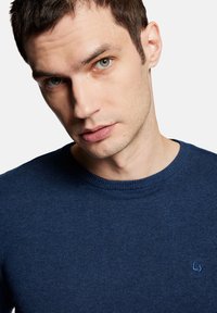 Granatowy sweter dzianinowy z okrągłym dekoltem, z małym niebieskim logo na lewej piersi i ściągaczami na mankietach.