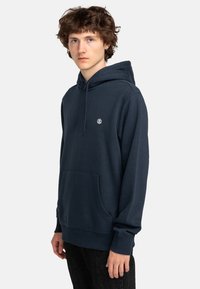 Navyblauwe hoodie van zacht stof, met een voorkant kangaroo zak en een verstelbare trekkoordcapuchon. Klein logo op de borst.