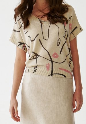 Kobieta w beżowym dzianinowym topie z abstrakcyjną czarno-różową grafiką linii, połączonym z jasnobeżową spódnicą, stojąca na białym tle.