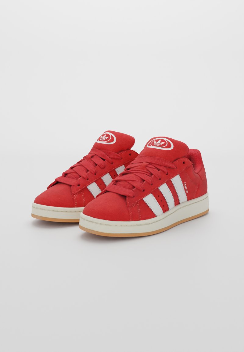 Czerwone zamszowe sneakersy z kontrastującymi białymi paskami, okrągłym logo na języku i beżową gumową podeszwą. Posiadają sznurówki oraz perforowane akcenty.