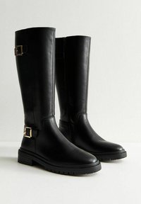 Un par de botas largas de cuero negro con suelas gruesas y detalles de hebilla dorada cerca del tobillo y la parte superior de la pantorrilla, apoyadas sobre una superficie blanca.