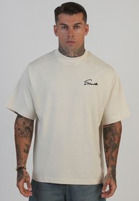 Beige oversized T-shirt i blød bomuld med korte ærmer, rund halsudskæring og et sort skriftlogo på venstre bryst.
