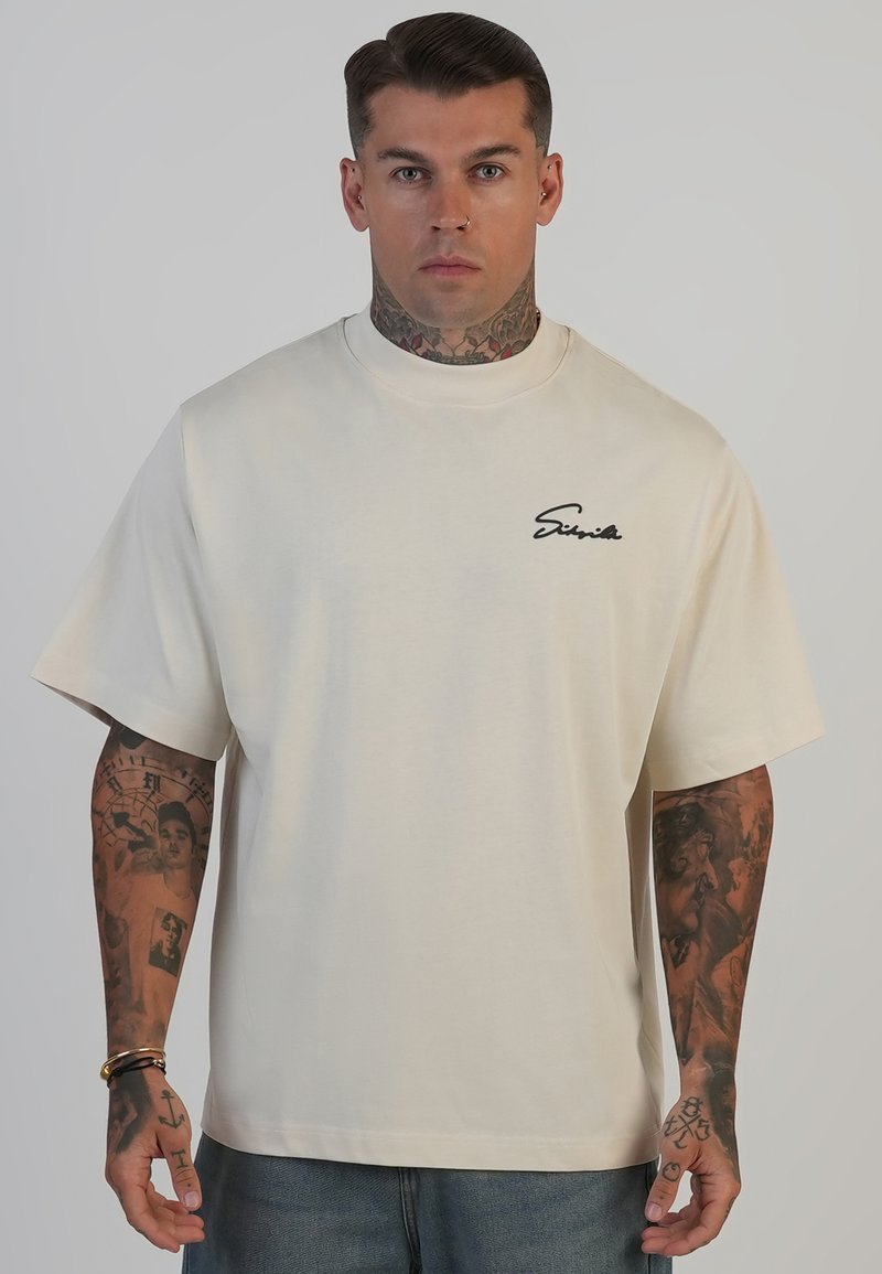 Beige oversized T-shirt i blød bomuld med korte ærmer, rund halsudskæring og et sort skriftlogo på venstre bryst.