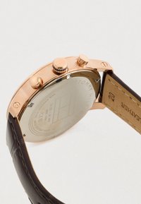 Retro della un orologio da polso in oro rosa con due pulsanti, una corona e un cinturino in pelle marrone che mostra dettagli delle cuciture e marcature.