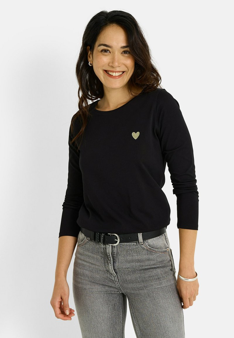 Camisa negra de manga larga con un acento dorado en forma de corazón, combinada con vaqueros grises y un cinturón negro. Tejido suave con un diseño ajustado.