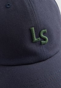 Casquette bleu marine en coton avec une texture lisse. Présente un logo "LS" brodé en vert à l'avant et des détails de couture visibles.