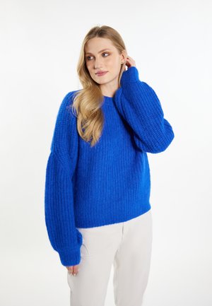 Maglione blu a maglia con una texture a coste, vestibilità oversized e maniche ampie, abbinato a pantaloni di colore chiaro.