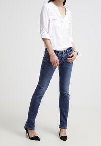 Blusa blanca con escote en V y dos bolsillos en el pecho, combinada con jeans ajustados de color azul oscuro y tacones altos negros. Textura plana, con detalles mínimos.