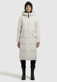 Lang hvit dunjakke med kviltet tekstur, frontglidelås, sidelommer og hette. Kombinert med svarte støvler og en strikket lue.