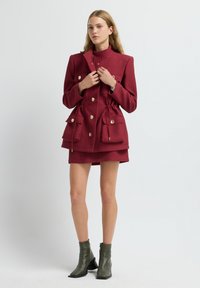 Cappotto rosso testurizzato con grandi tasche e bottoni dorati, abbinato a una gonna rossa coordinata. Il modello indossa stivali alla caviglia verdi.