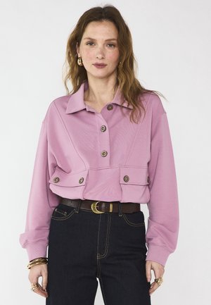 Femme aux longs cheveux roux portant une veste courte rose à boutons, un jean taille haute foncé, des boucles d'oreilles créoles en or ainsi que des bracelets et des bagues en or.