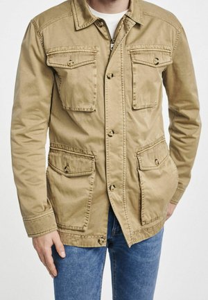 Mann trägt eine beige Knopfleistejacke mit vier Klapptaschen und blaue Jeans vor einfachem Hintergrund.